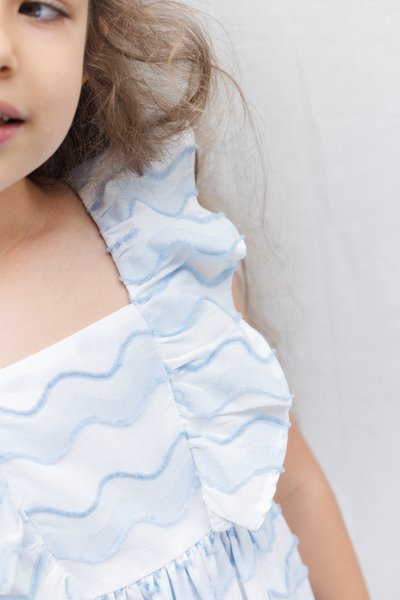 Λευκό φόρεμα με μπλε υφασμάτινες λουρίδες κυματιστού σχεδίου. Διαθέτει βολάν στους ιμάντες στους ώμους και πιέτες στη μέση. Ομαλή υφή.