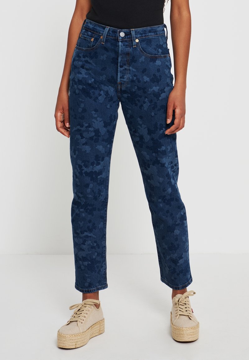 Personne portant un jean bleu taille haute à motif camouflage et des baskets beiges à plateforme, debout devant un fond blanc uni.