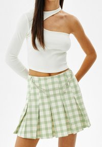 Vit ribbad crop top med en lång ärm och asymmetrisk halsringning, matchad med en grön rutiga veckad kjol med kedjedetaljer.