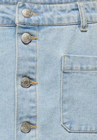 Tissu en denim bleu clair avec une rangée verticale de quatre boutons en métal et une poche plaquée carrée sur le côté droit.