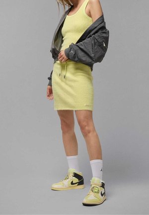 Kvinde iført lys gul ærmeløs kjole, grå jakke draperet over skuldrene, hvide sokker og gule og sorte Nike-sneakers stående på gråt gulv.