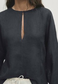 Blouse en lin bleu marine foncé avec un col rond, une ouverture en forme de goutte d'eau, et des manches longues. Elle présente une coupe ample et un cordon de serrage ajustable à l'ourlet.