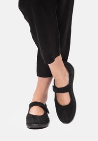 Schwarze Wildleder-Mary-Jane-Schuhe mit runder Zehenpartie, einem gewellten Kantendesign und einem Klettverschlussriemen. Auf einem hellen Hintergrund platziert.