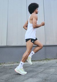 Man with afro hairstyle running outdoors wearing white tank top, black shorts, white socks, and light green running shoes.