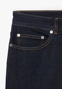 Vaqueros de mezclilla oscura con una textura suave, costuras en contraste de color naranja y herrajes plateados. Incluye un bolsillo frontal con remaches.