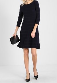 Vestido a rayas azul marino con mangas tres cuartos, falda acampanada, hecho de tela texturizada. Combinado con un pequeño bolso negro y tacones altos.