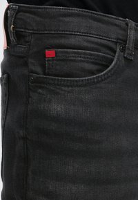 Nærbillede af sort denimjeanslomme med et lille rødt stofmærke med teksten "HUGO" på møntlommen.