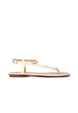 Flad beige lædersandal med knyttet rem og justerbar ankelspænde på en tynd brun sål.