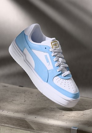 Weißer und hellblauer Puma-Sneaker mit perforierter Spitze, weißen Schnürsenkeln und goldakzentuiertem Logo, der über einer Betonoberfläche schwebt.