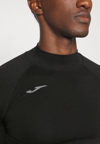 Schwarzes, athletisches Langarmshirt aus strukturiertem Material. Besitzt einen hohen Kragen und ein kleines graues Logo auf der Brust. Glattes und tailliertes Design.