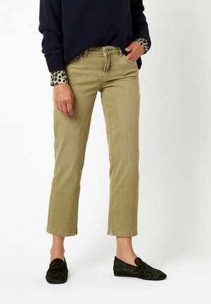 Person trägt beige gerade geschnittene Hose, schwarze gemusterte Loafers, dunkelblauen Pullover und sichtbare Manschetten eines Leopardenmuster-Hemds.