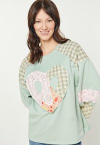 Világos zöld kapucnis pulóver, nagy rózsaszín szív applikációval és mintás anyagokkal. Kockás díszítések a ujjakon és a vállakon.