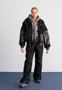 Zwarte bomberjack met bontkraag, gebloemde hoodie eronder, zwarte wijde broek en donkere sneakers. Rood labelaccent op het jack.