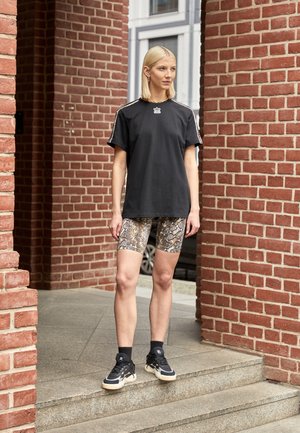 Sort grafisk t-shirt med korte ærmer, parret med cykelshorts med slangehudsmønster. Sorte sneakers med hvide og brune nuancer. Murstensbaggrund.