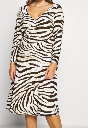 Žena v dlouhých rukávech a zavinovacích šatech po kolena s černobílým zebřím vzorem stojí před jednobarevným pozadím.