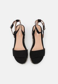 Sandali in suede neri con una larga fascia frontale, cinturino alla caviglia con fibbia e suola in stile espadrilles intrecciato. Design piatto e aperto per la traspirabilità.
