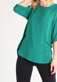 Blusa verde, leggera, con scollatura rotonda e maniche a tre quarti. Presenta una vestibilità ampia e un'orlo curvo. Tessuto morbido.