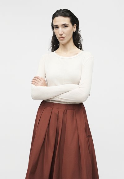 Γυναίκα με μακριά σκούρα μαλλιά, φορώντας λευκό μακρυμάνικο τοπ και σκουρόχρωμη φούστα με πιέτες, στέκεται με σταυρωμένα τα χέρια πάνω σε λευκό φόντο.