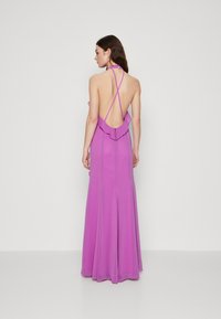 Robe de soirée longue rose avec un col halter, un dos ouvert avec des sangles croisées, des accents volants au niveau du décolleté, et une jupe fluide.