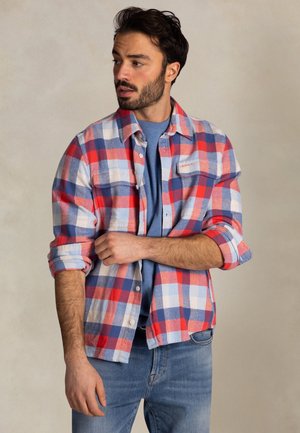 Baardige man die de mouw aanpast van een blauw, rood en wit geruit overhemd dat gedragen wordt over een blauw T-shirt en een spijkerbroek, terwijl hij naar links kijkt.