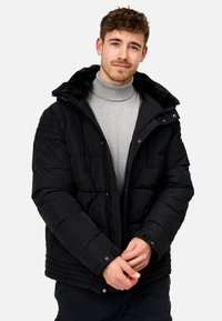 Schwarze Pufferjacke mit gefütterter Kapuze aus Kunstfell, Reißverschluss und Druckknopfverschlüssen, gepolstertem Design und geripptem Saum. Getragen über einem grauen Rollkragenpullover.