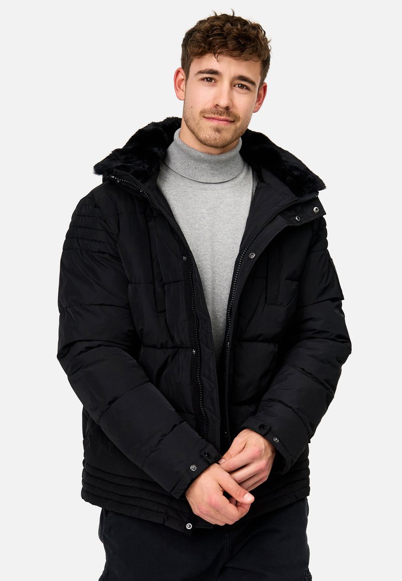 Schwarze Pufferjacke mit gefütterter Kapuze aus Kunstfell, Reißverschluss und Druckknopfverschlüssen, gepolstertem Design und geripptem Saum. Getragen über einem grauen Rollkragenpullover.