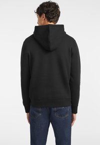 Fekete kapucnis pulóver, állítható kapucnival, laza szabású, bordázott mandzsettákkal. Farmerjeans-szel párosítva, amelyeken hátsó zsebek vannak, és egy finom kék textúra.