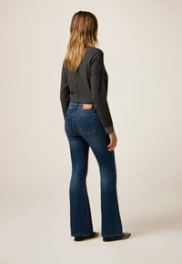 Maglione grigio scuro con bottoni sulla schiena, abbinato a jeans blu svasati con etichetta in pelle marrone in vita e polsini decorati.