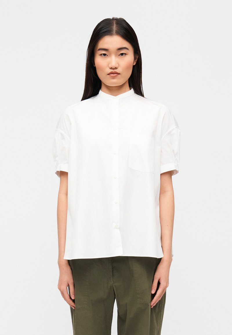 Άσπρο πουκάμισο με κοντά μανίκια και μανδύα μανδ αρίνου, με μπροστινή σειρά κουμπιών και μια τσέπη στο στήθος. Λείο βαμβακερό ύφασμα.