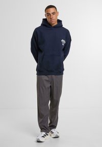 Tamsiai mėlynas džemperis su kapišonu, su priekyje esančia kišenėle ir mažuoju baltu logotipu, derinamas su pilkais kelnais ir baltais sportbačiais su juodomis juostelėmis.