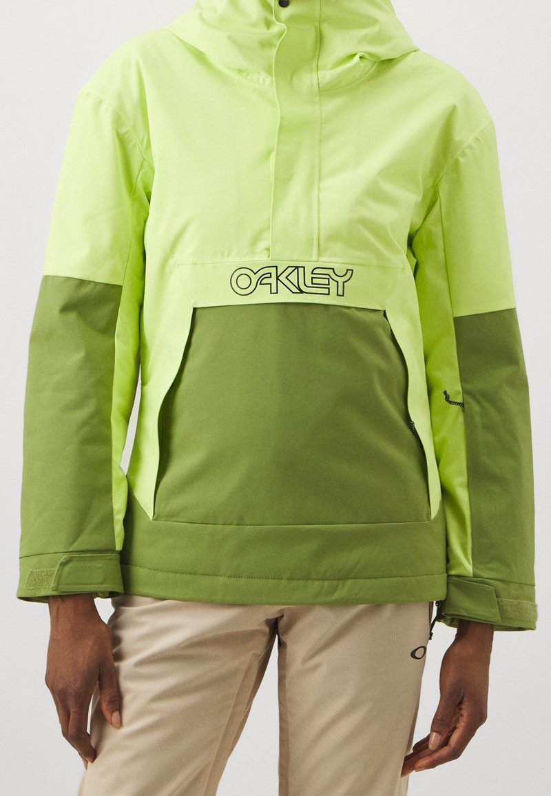 Personne portant une veste à capuche Oakley verte et vert clair avec un pantalon beige, les mains le long du corps, debout devant un fond uni.