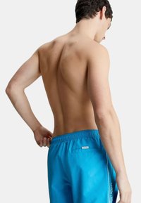 Pantaloni da bagno blu realizzati in tessuto leggero, dotati di una cintura elastica, un patch con logo e piping laterale a contrasto. Vista dal retro.
