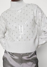 Pull blanc en laine tricotée avec un col montant, orné de paillettes et de perles argentées disposées de manière éparse. L'ourlet côtelé ajoute de la texture.