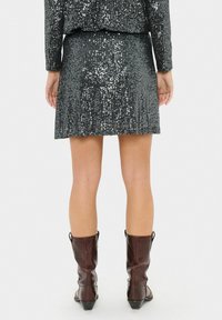 Mujer vestida con una falda y un top de lentejuelas negras, combinados con botas de cuero marrón a media pantorrilla, de pie de espaldas sobre un fondo blanco.