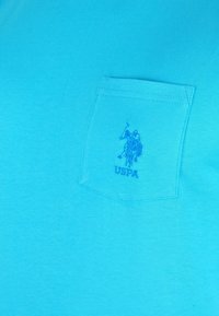 Turkos t-shirt med kort ärm i bomull med en vänster bröstficka. Har en broderad logotyp i blått som föreställer en polo-spelare.