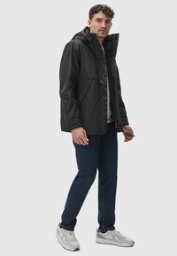 Zwarte waterdichte jas met een capuchon, rits aan de voorkant en zijzakken, gecombineerd met donkerblauwe jeans en wit-grijze sneakers.