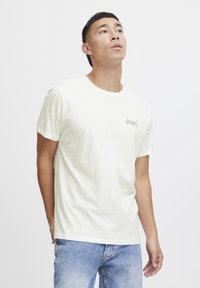 White short-sleeve t-shirt made of soft fabric featuring a small green logo on the left chest. Worn with light blue jeans.