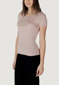 Camiseta de manga corta rosa claro hecha de tela suave, que presenta un logo metálico "EA7 EMPORIO ARMANI" en la parte delantera con detalles en dorado.