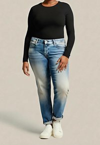 Personne portant un jean bleu clair délavé, un t-shirt noir à manches longues et des baskets blanches, debout sur un fond neutre.