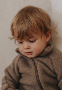 Otrok v rjavi flis jakni z zadrgo, z visokim ovratnikom in gladko teksturo, sedi ob nevtralnem ozadju, osredotočen na nekaj spodaj.