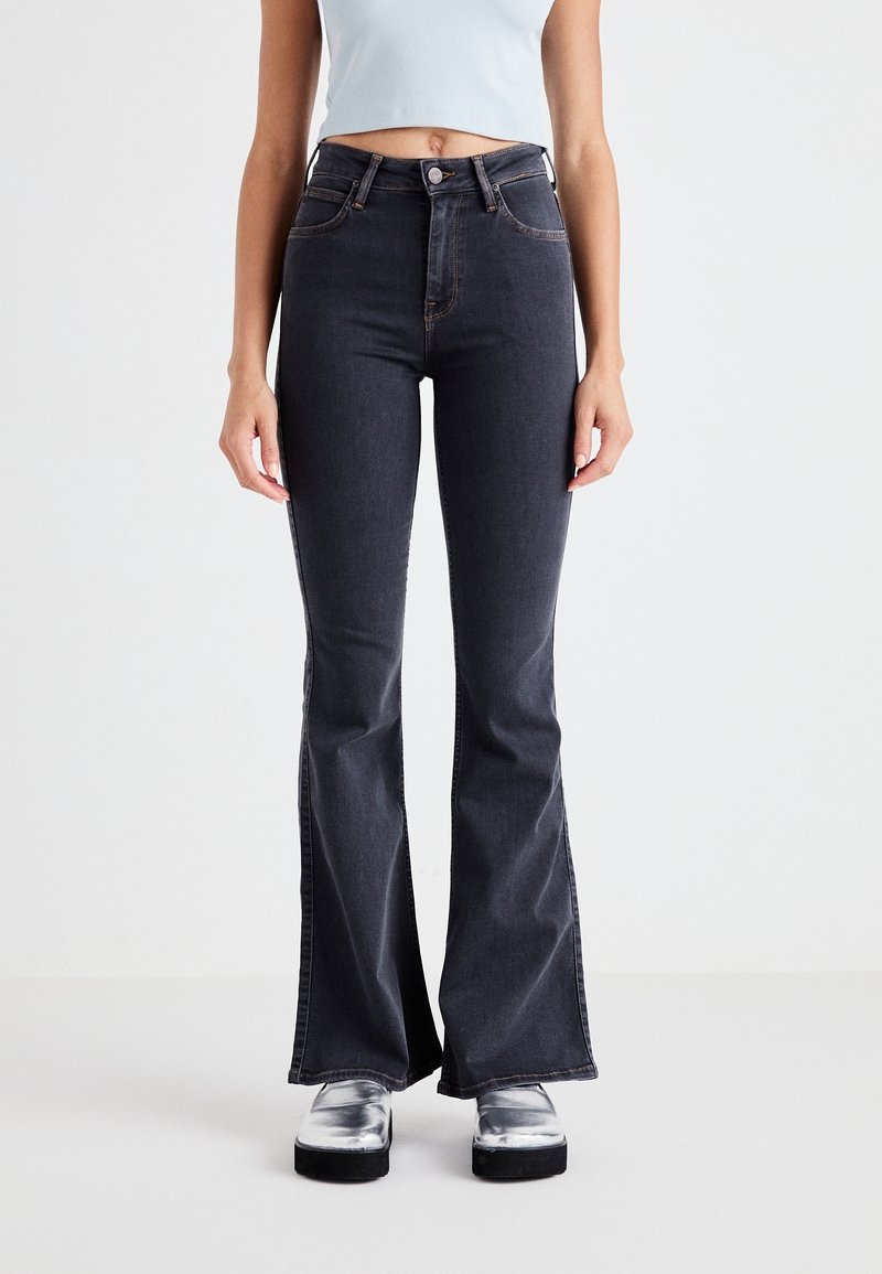 Femme portant un jean flare taille haute gris foncé, associé à des chaussures plateformes argentées et un débardeur court bleu clair sur un fond uni.