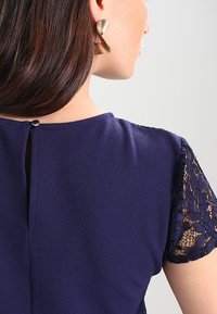 Abito blu navy con maniche corte in pizzo, chiusura posteriore a occhiello e texture liscia, abbinato a orecchini triangolari dorati.