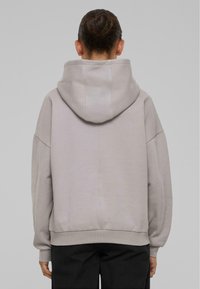 Lichtgrijze hoodie met een grote capuchon, verlaagde schouders en een geribbelde zoom. Materiaal lijkt zacht met een gladde textuur; minimalistisch ontwerp.