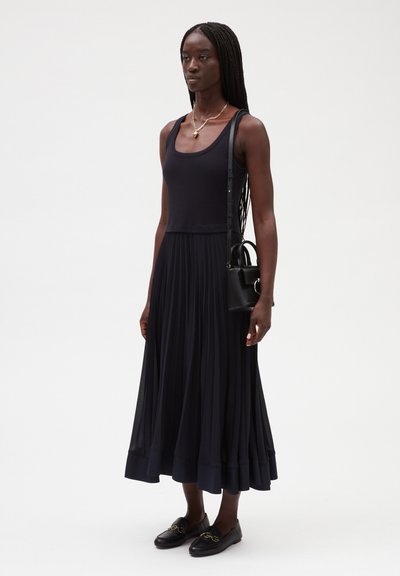 Vestido largo plisado de color navy con un corsé de top ajustado, falda fluida, combinado con un pequeño bolso negro y mocasines con detalles dorados.
