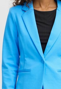 Kvinna i en klarblå kavaj över en svart ribbad topp, visar överkroppen samt kavajens knapp och slag.