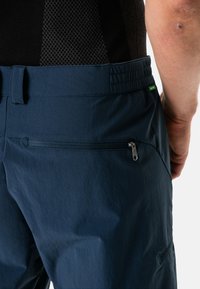 Pantalones azul marino con cintura elástica y bolsillo lateral con cremallera. El material parece ligero con una textura suave. Presenta un pequeño logo verde.