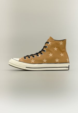 Høytgått brun canvassneaker med lyst stjernemønster, svarte lisser, hvit gummisåle og svart stripedetalj på nøytral bakgrunn.