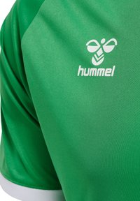 Grön atletisk tröja i glatt tyg, med en vit "hummel"-logotyp på bröstet och en enkel vit kant vid kragen.