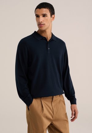 Jeune homme aux cheveux courts et bouclés, portant un polo à manches longues bleu marine foncé et un pantalon beige, debout avec une main dans la poche.