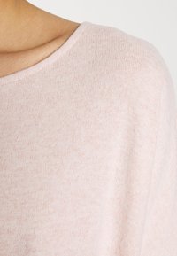 Pull en tricot rose clair avec un large col et une texture douce, présentant un motif chiné subtil sur toute la surface.